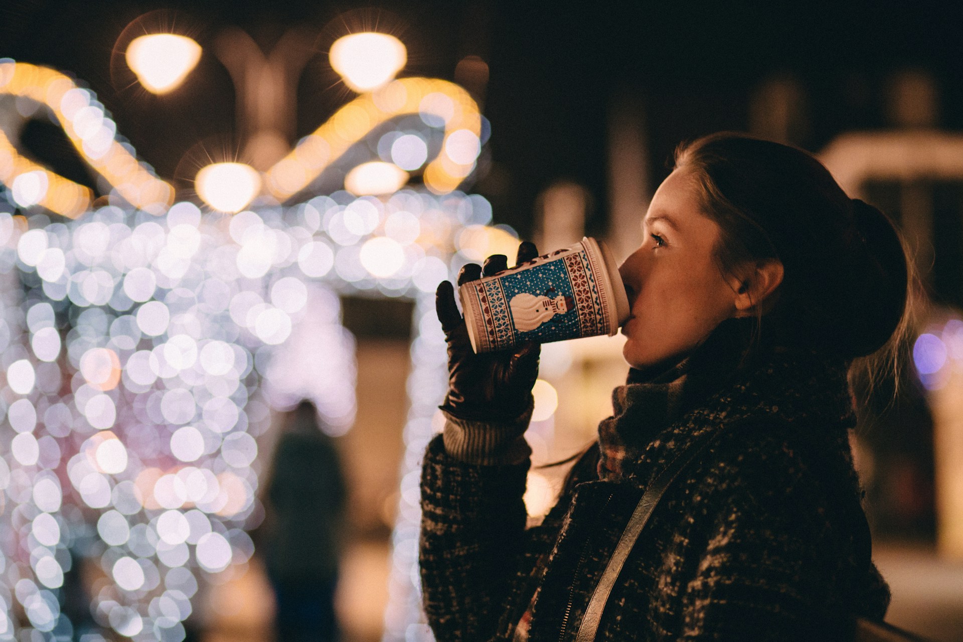 Woman drinking hot cocoa out of a snowman cup while looking at holiday lights.